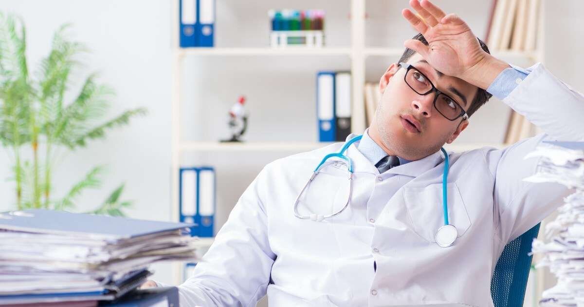 young male doctor sitting at desk seeming stressed with hand on forehead 